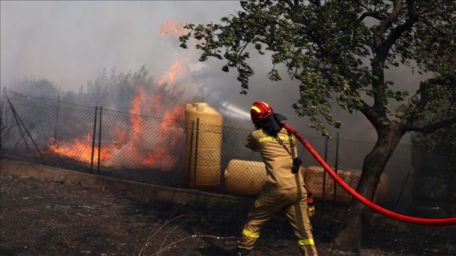 Aerial view of a massive wildfire burning through a forested area in Attica, Greece, with thick smoke rising into the sky.