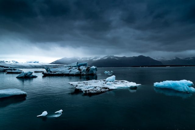 A massive Antarctic ice sheet stretches across the horizon under a pale blue sky, with penguins gathered on a rocky outcrop in the foreground.