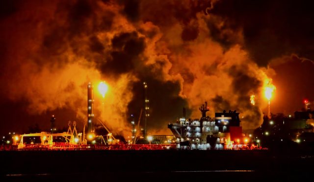 Oil barrels stacked at a storage facility with a price ticker displaying Brent Crude and West Texas Intermediate values.