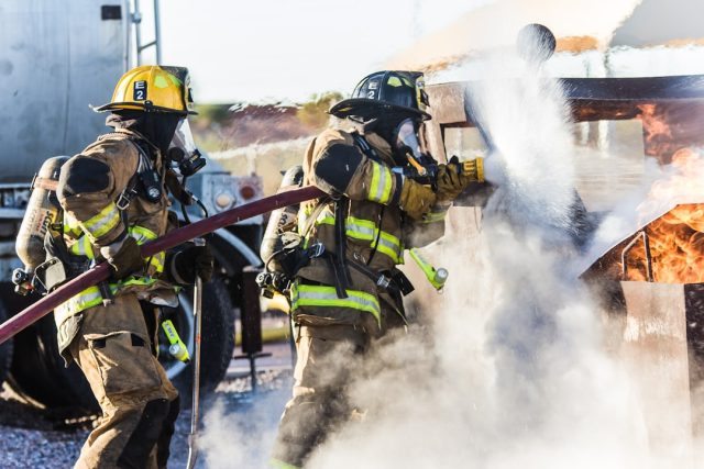 Firefighter in full gear stands near a fire truck, with a smoke-filled sky in the background.