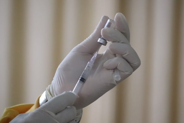 A nurse holds a measles vaccine vial beside a calendar marked 12-15 months, symbolizing the infant vaccination window.