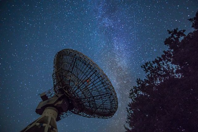 A radio telescope dish points toward the night sky, capturing a bright burst of light from a distant molecular cloud.