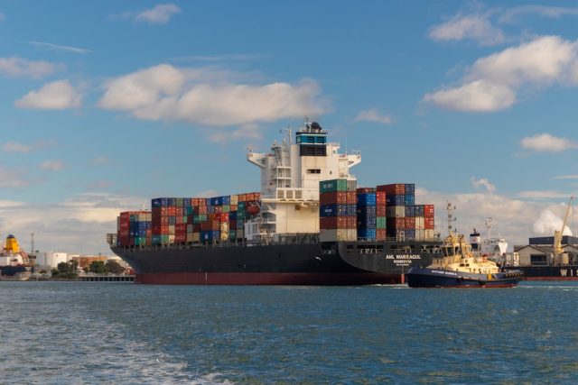A cargo ship navigates the narrow Bab el-Mandeb strait, with military vessels visible on the horizon.