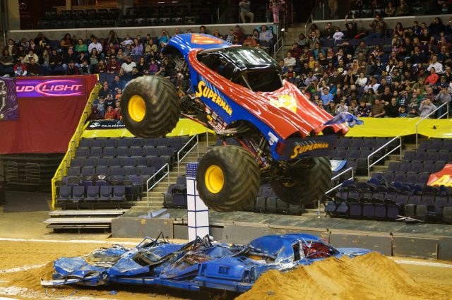 A large monster truck with oversized tires sits on a dirt track as spectators watch from behind barriers at an exhibition event.