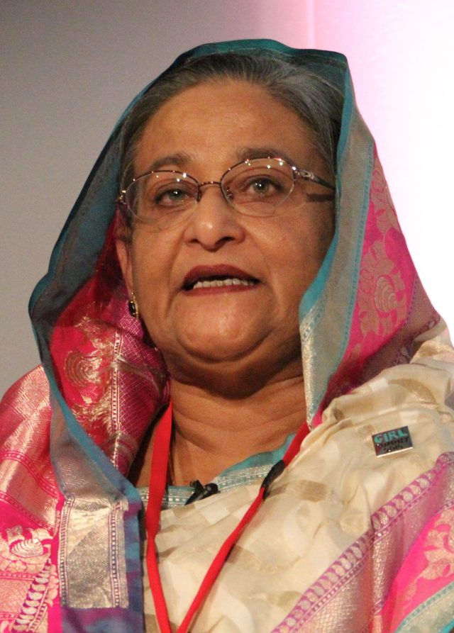Sheikh Hasina speaking at a podium with Bangladesh flag in background during a political event.