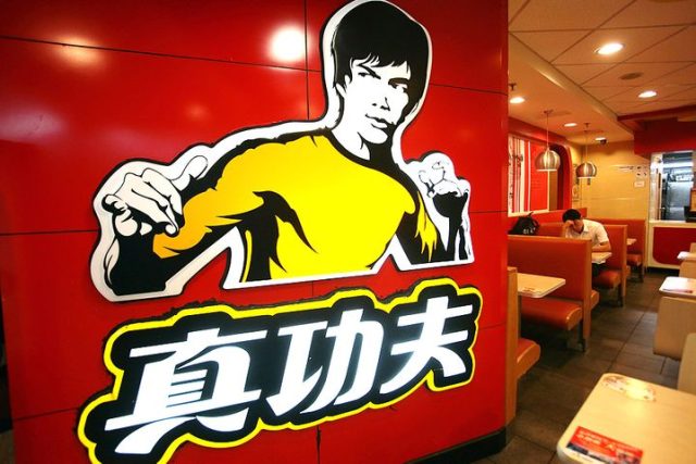 Real Kungfu Bruce Lee Logo Shannon Lee stands at a podium holding a photo of her father Bruce Lee beside the Real Kungfu restaurant logo for comparison.