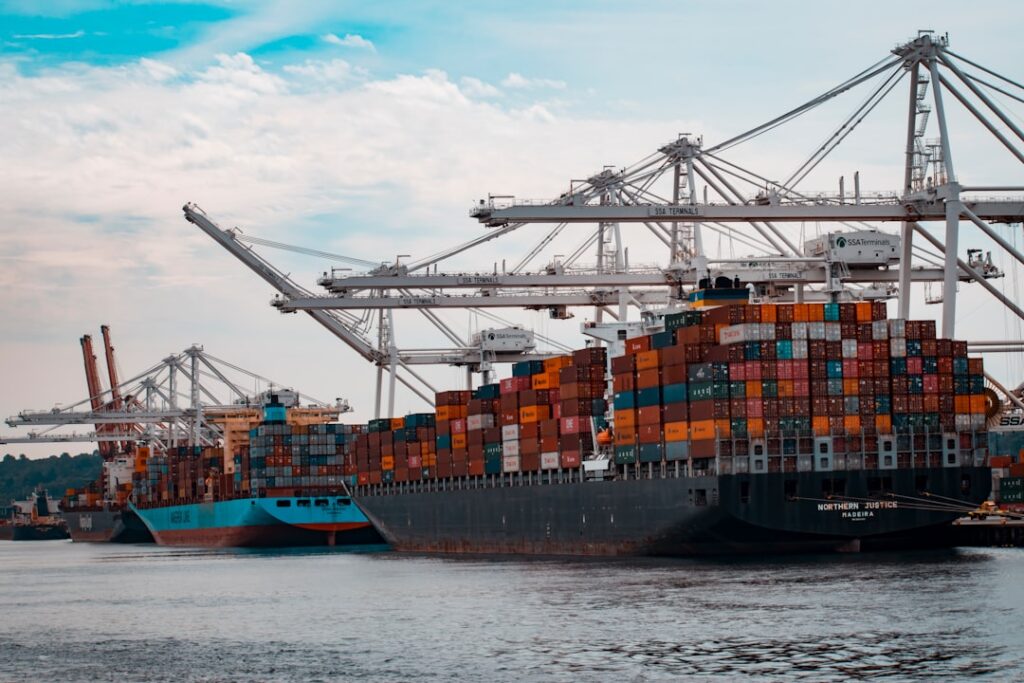 Cargo containers stacked at a Malaysian port, with a ship loading goods under a clear sky.